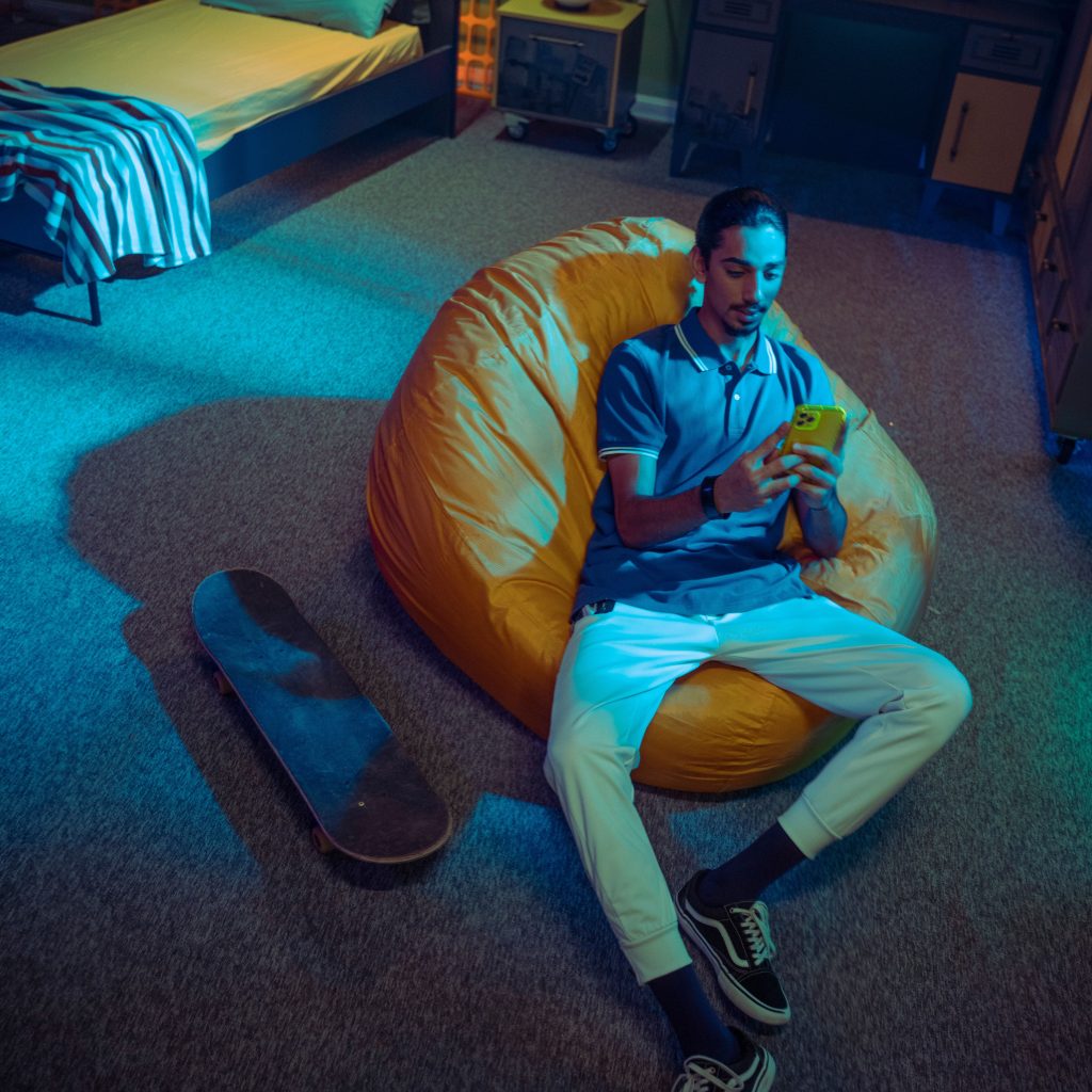 Un giovane uomo in una stanza elegante controlla lo smartphone mentre è seduto su un pouf accanto a uno skateboard.